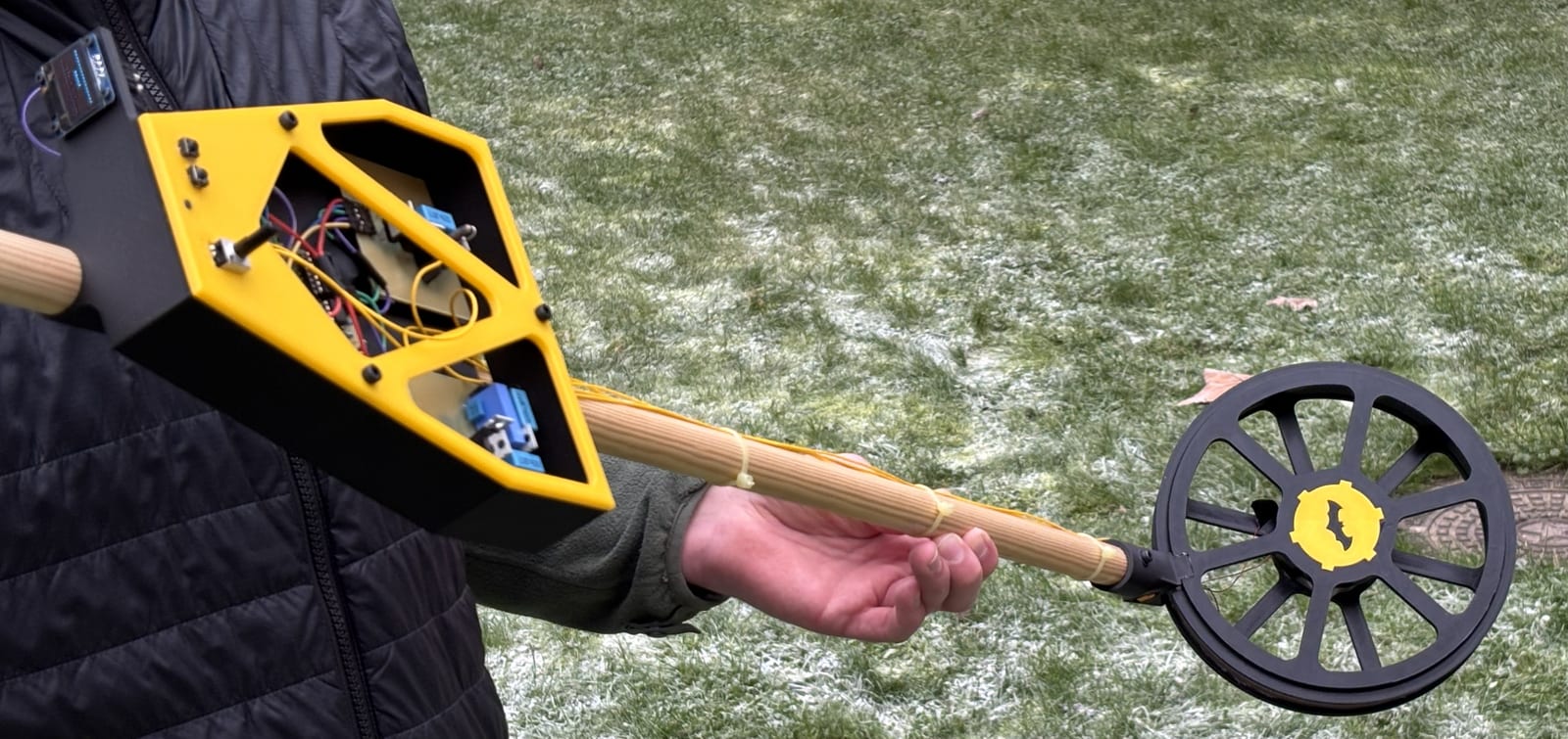 The finished metal detector held outdoors — yellow 3D-printed control housing with visible electronics, wooden shaft, and concentric coil head on frosty grass